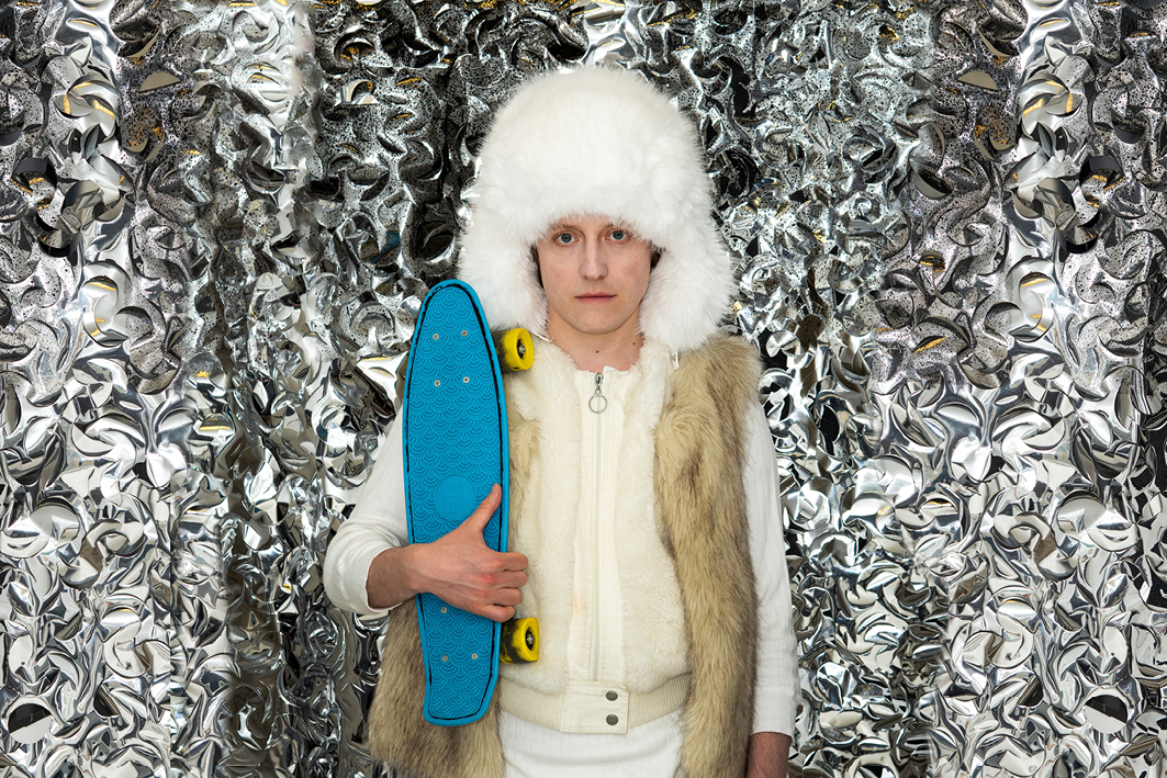 Person mit Pelzmütze und Pelzmantel steht vor glitzerndem Vorhang und hat ein Skateboard im Arm.